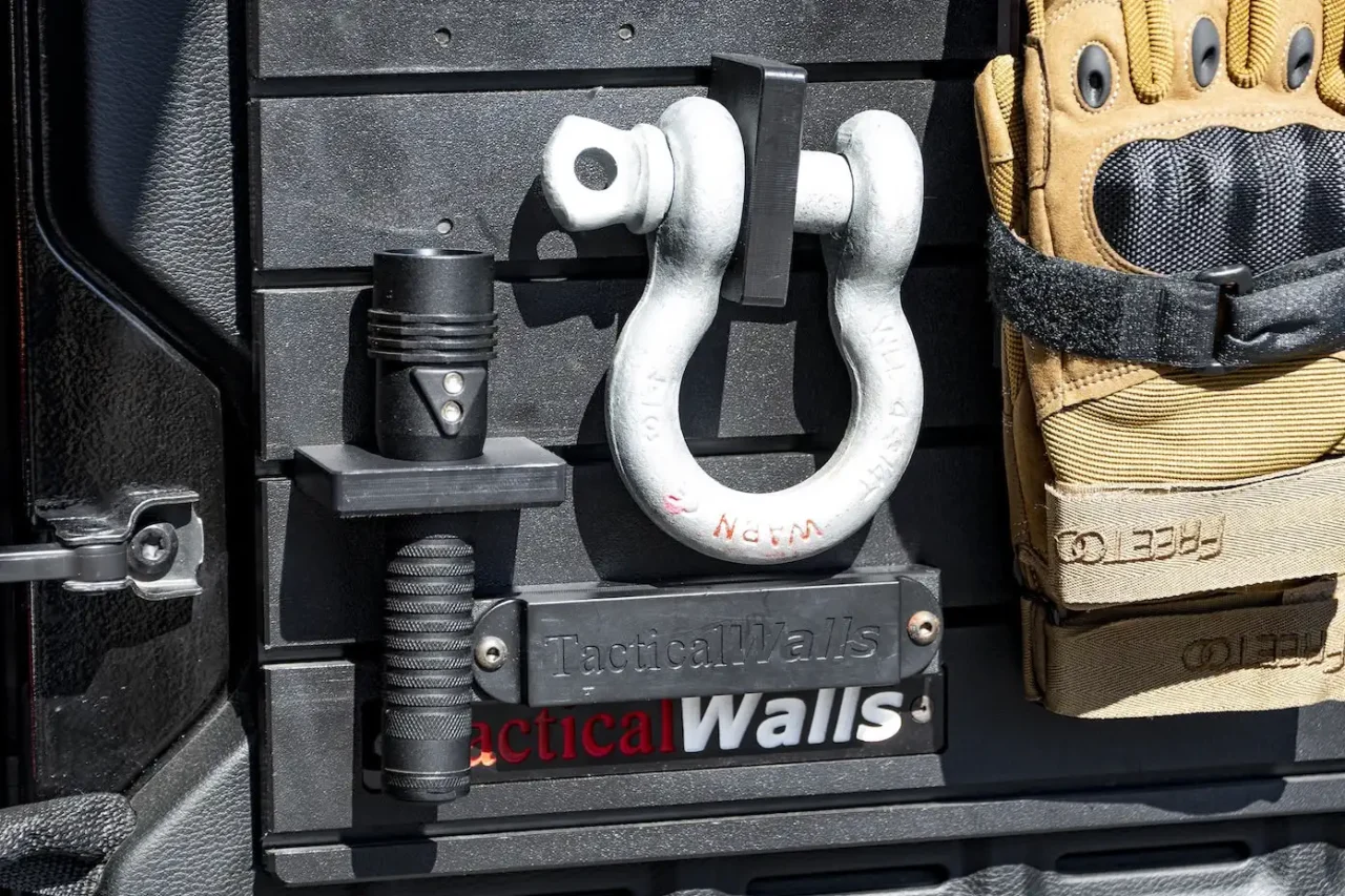 ModWall Flashlight Holder A black handheld flashlight secured vertically in the ModWall Flashlight Holder on a gear-loaded tailgate panel, organized alongside a recovery shackle on the flashlight holder and work gloves.