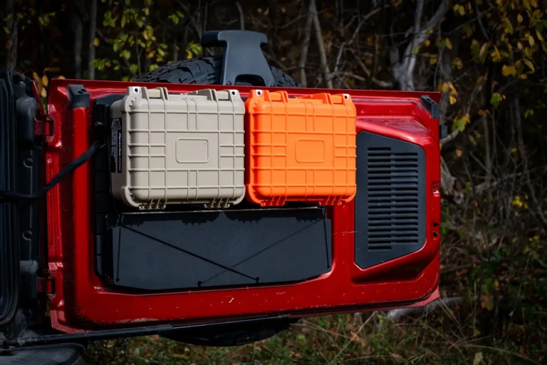 An open red Ford Bronco tailgate featuring the modular storage package with two hard-shell gear cases (one orange, one tan) and a folded tailgate table mounted to the black tailgate panel.